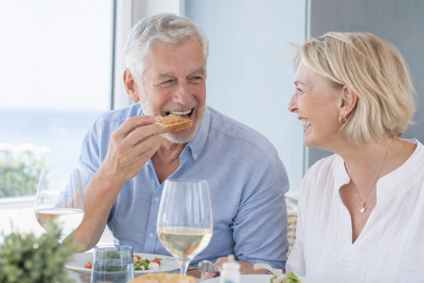 A couple in their sixties laughing over dinner as the man bites into crusty bread